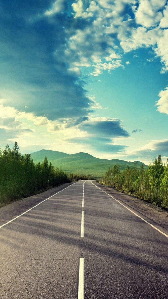 An asphalt road stretches toward green hills under a blue sky with white clouds