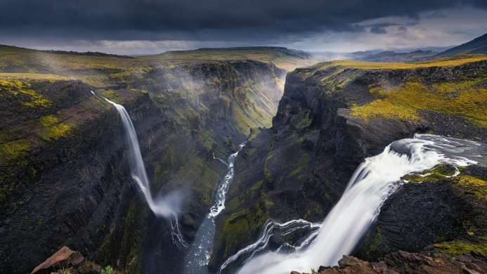 A dramatic landscape with a canyon and two waterfalls, under a dark cloudy sky