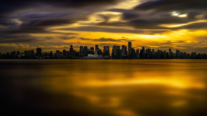 City skyline reflecting on water at sunset with a dramatic sky, gold and black tones