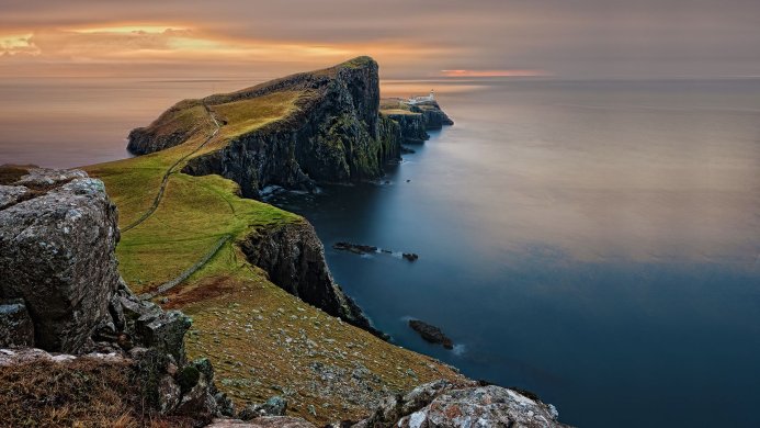 Scenic view of a grassy cliff and lighthouse on a coastline under an orange sky