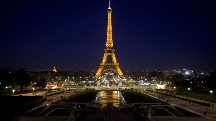 Eiffel Tower at night, illuminated against a dark blue sky, with city lights in the background