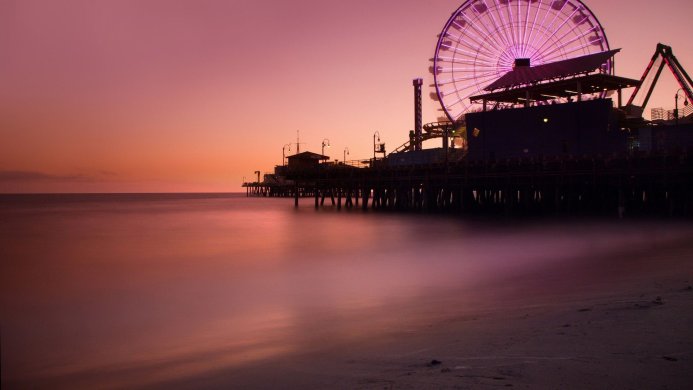 Ferris wheel and pier at sunset with a smooth water surface in shades of purple and orange