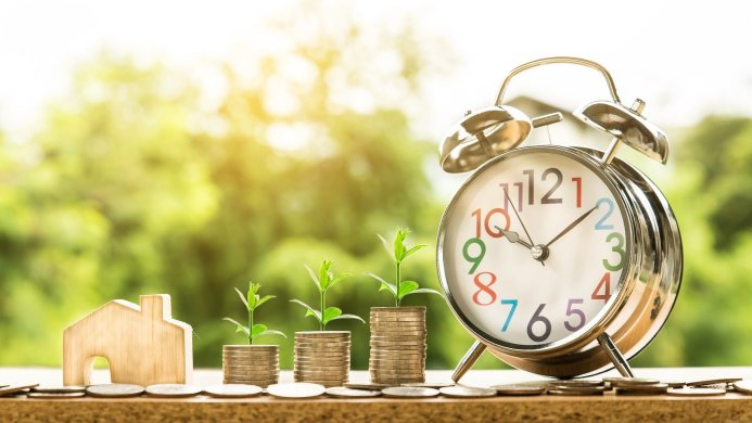 Alarm clock, coin stacks with seedlings, and a wooden house representing financial growth