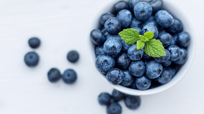 A bowl of fresh blueberries garnished with a sprig of mint on a white surface