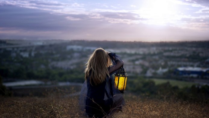 A girl holding a lantern, looking out over a landscape at dusk