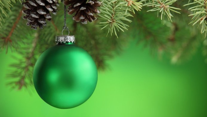 A green Christmas ornament hangs from a pine branch with pine cones on a green background