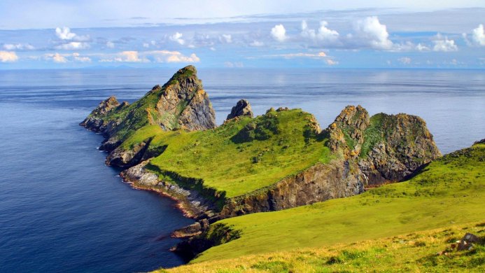 Scenic view of a green island with rocky cliffs surrounded by blue ocean and a cloudy sky