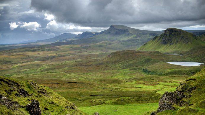 A scenic view of a valley with green hills and mountains under a cloudy sky