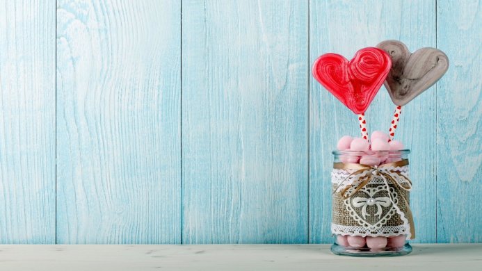 Two heart lollipops in a jar of pink marshmallows, tied with lace and ribbon on a blue wooden background