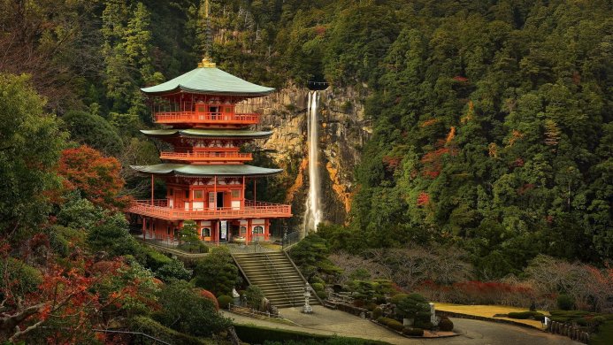 A red and green pagoda stands nestled in a lush green forest with a waterfall in the background