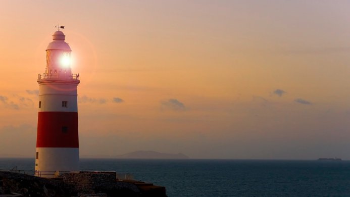 A lighthouse with a bright beacon against an orange sunset sky by the sea