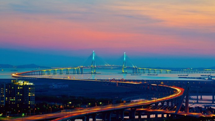 A long bridge spans a body of water under a colorful sky during twilight