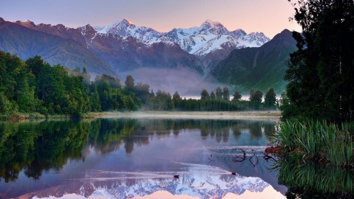 Lake reflecting snow-capped mountains and forest at dawn or dusk