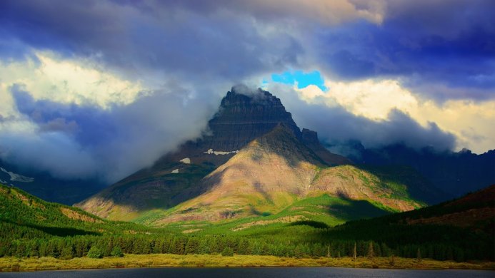 A mountain peak surrounded by clouds, partially lit by sunlight, with a forest and lake in the foreground