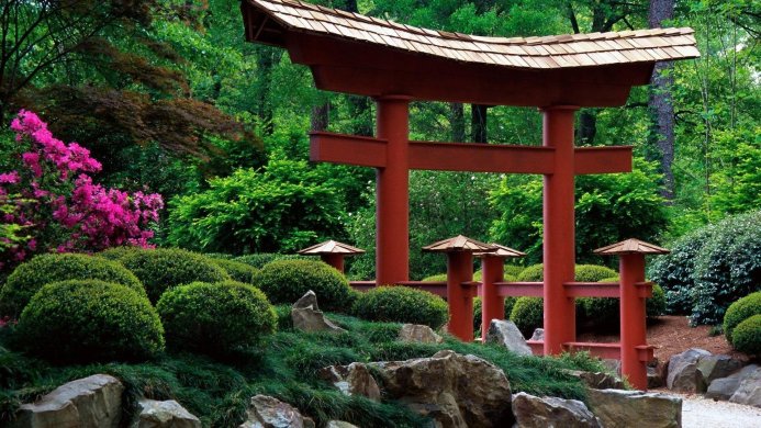 A red Torii gate stands in a lush green Japanese garden with rocks and pink flowers
