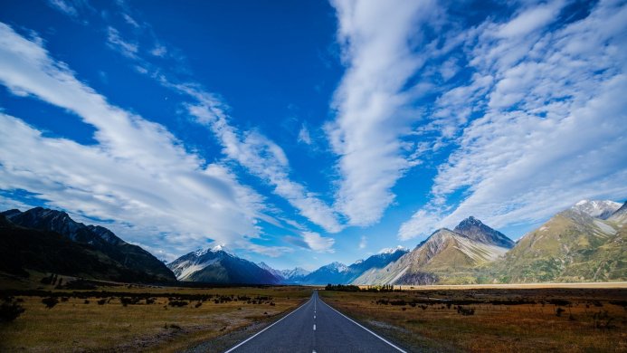 A long road stretching towards snow-capped mountains under a bright blue sky with scattered clouds