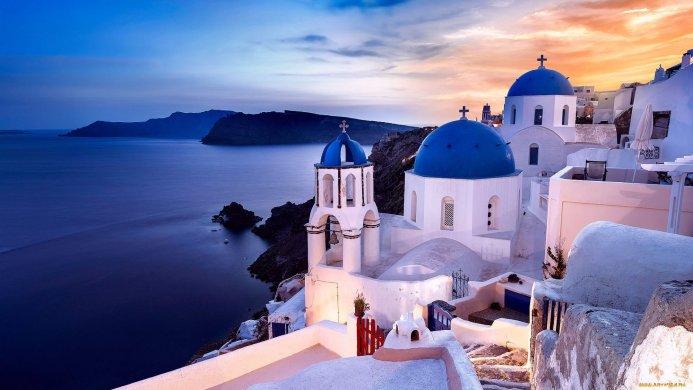 White buildings with blue domes on a cliff overlooking the sea at sunset