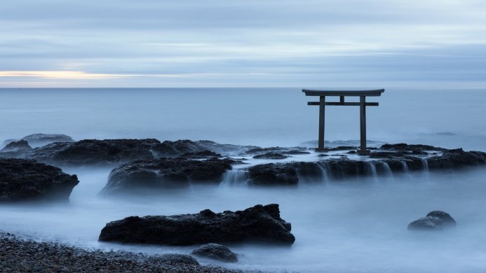 A torii gate standing on rocks in the ocean with a misty, dreamlike effect