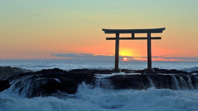 A torii gate on a rocky shore at sunrise, with ocean waves crashing