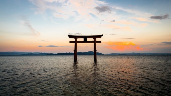 A torii gate stands in water at sunset with mountains in the distance