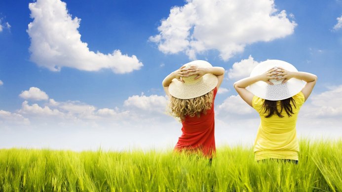 Two women wearing straw hats in a green field under a blue sky with white clouds
