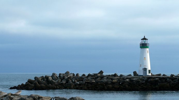 A white lighthouse stands on a rocky pier with the ocean and a cloudy sky in the background