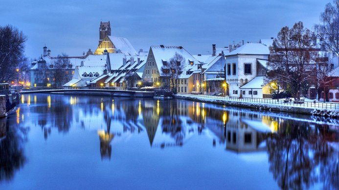 Winter cityscape with snow-covered buildings reflecting in a river during twilight hours