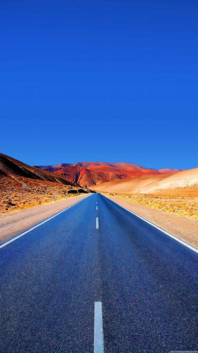 Blue road leading to brown and red mountains under a clear blue sky