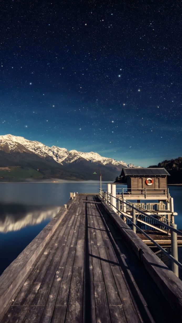 A wooden dock on a calm lake with snow-capped mountains in the background under a starry night sky