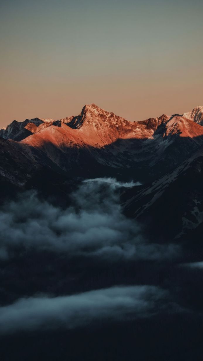 Snowy mountain peaks rise above a sea of clouds under a dusky orange sky
