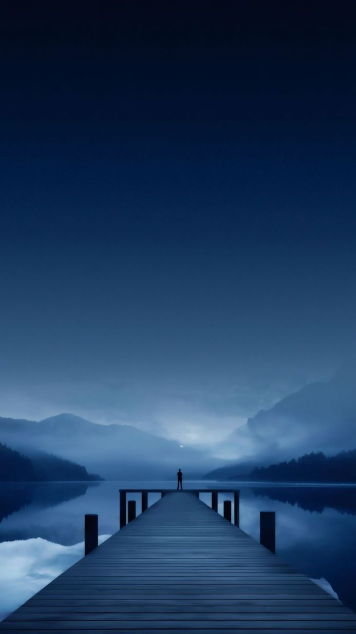 Silhouette of a person standing on a pier overlooking a lake and mountains in blue tones