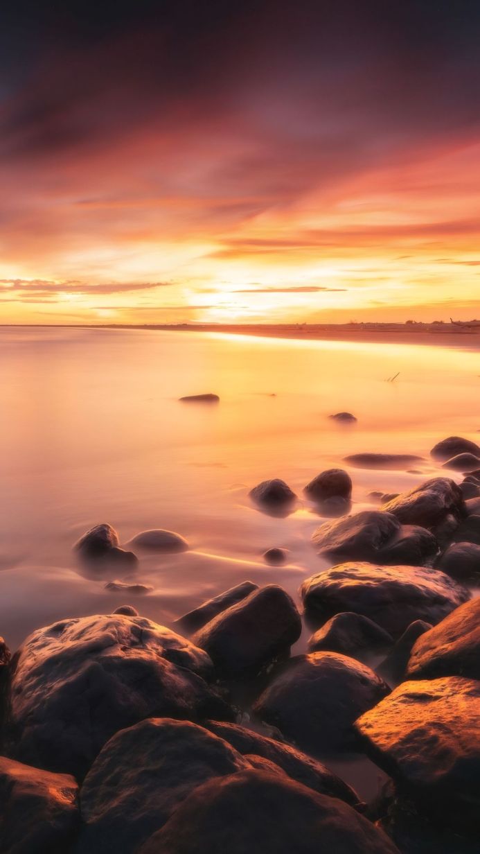 A seascape with rocks in the foreground, smooth water and a colourful sky at sunset