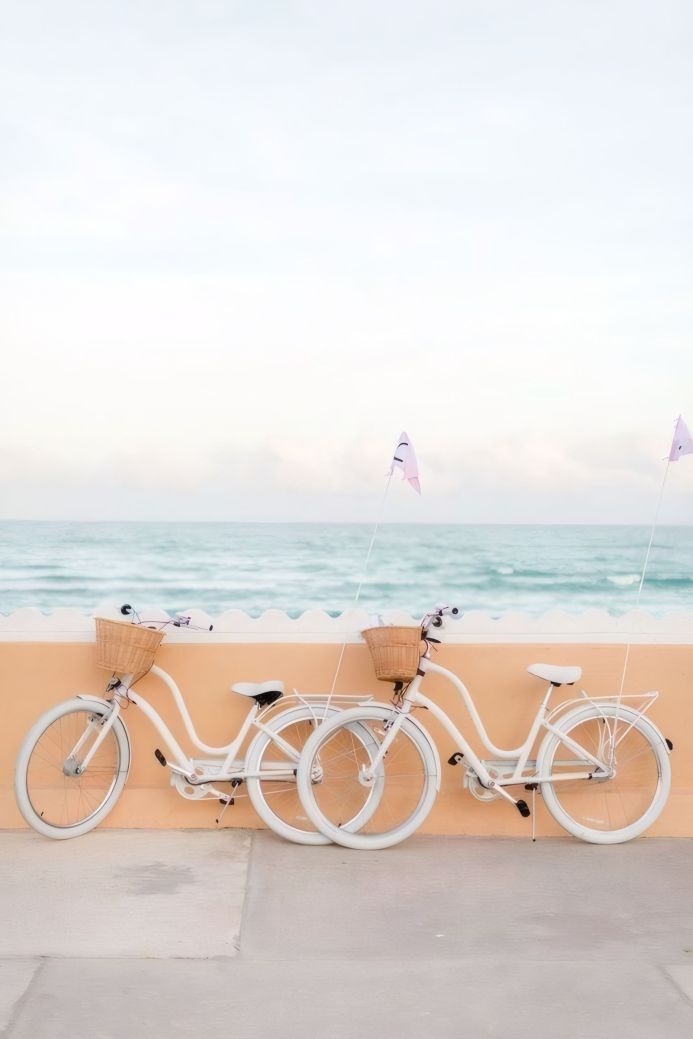 Two white bicycles with baskets parked by the beach with pink flags