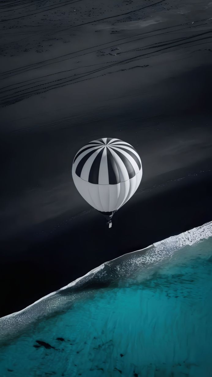 A black and white hot air balloon over turquoise ocean water and a black sand beach